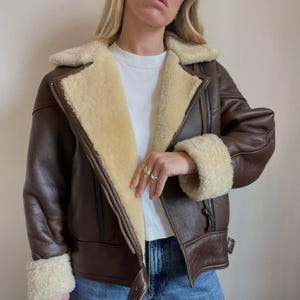 May include: A brown leather jacket with a cream-colored shearling lining and trim. The jacket features a zip closure, a wide collar, and shearling cuffs. The jacket is a classic aviator style.
