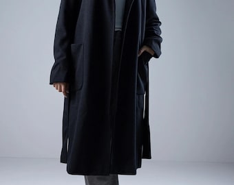 Puede incluir: Un abrigo largo de color gris oscuro con capucha y bolsillos. El abrigo está abierto por delante y tiene un cinturón a juego. La modelo lleva pantalones grises y zapatos negros. El abrigo parece estar hecho de un material suave y cálido.