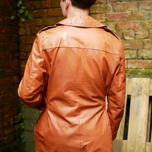May include: A vintage brown leather jacket with a collared neckline and button closure. The jacket has a tailored fit and is worn over blue jeans. The jacket's length extends to the upper thighs. The background features a brick wall and wooden planks.