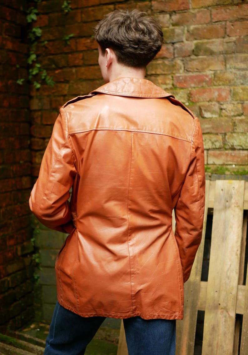 May include: A vintage brown leather jacket with a collared neckline and button closure. The jacket has a tailored fit and is worn over blue jeans. The jacket's length extends to the upper thighs. The background features a brick wall and wooden planks.