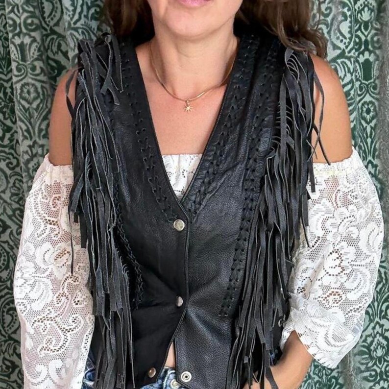 May include: Black leather vest with fringe detailing. The vest has a V-neck and is buttoned down the front. The sleeves are cut off, and the vest is worn over a white lace top. A gold necklace with a small pendant is visible.