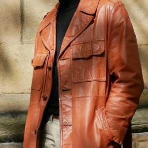 May include: A brown leather jacket with multiple pockets and a collared neckline. The jacket has a button-up front and is worn over a black turtleneck. The jacket appears to be vintage or retro style.