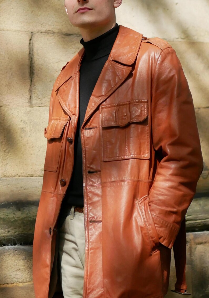 May include: A brown leather jacket with multiple pockets and a collared neckline. The jacket has a button-up front and is worn over a black turtleneck. The jacket appears to be vintage or retro style.