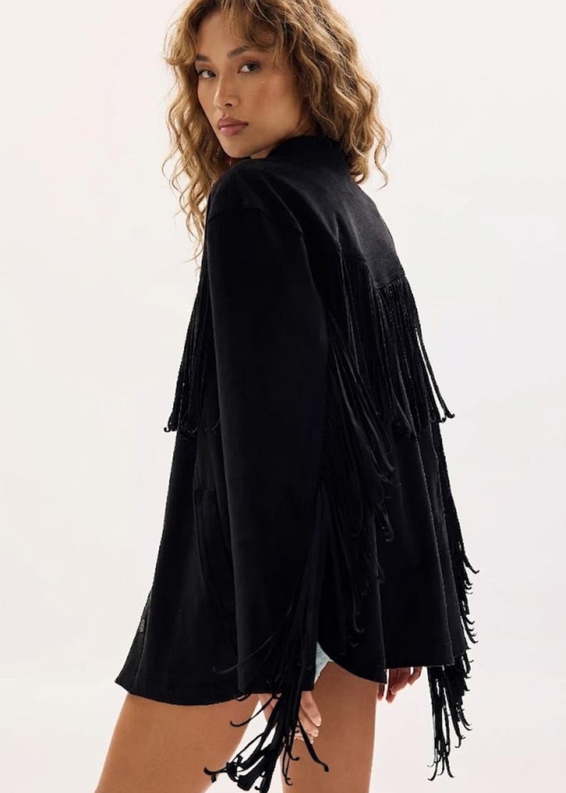 May include: Black jacket with long fringe detailing. The jacket has a high collar and a loose fit. The fringe is long and hangs down from the back and sleeves. The jacket is made of a soft, flowing fabric.