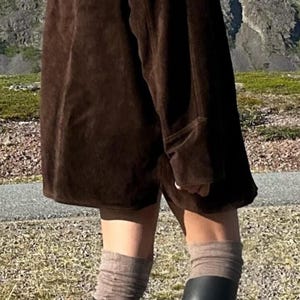 May include: A person wearing a dark brown suede jacket and matching shorts. The outfit is paired with gray socks and black rubber boots. The background features a mountain range and a paved path.
