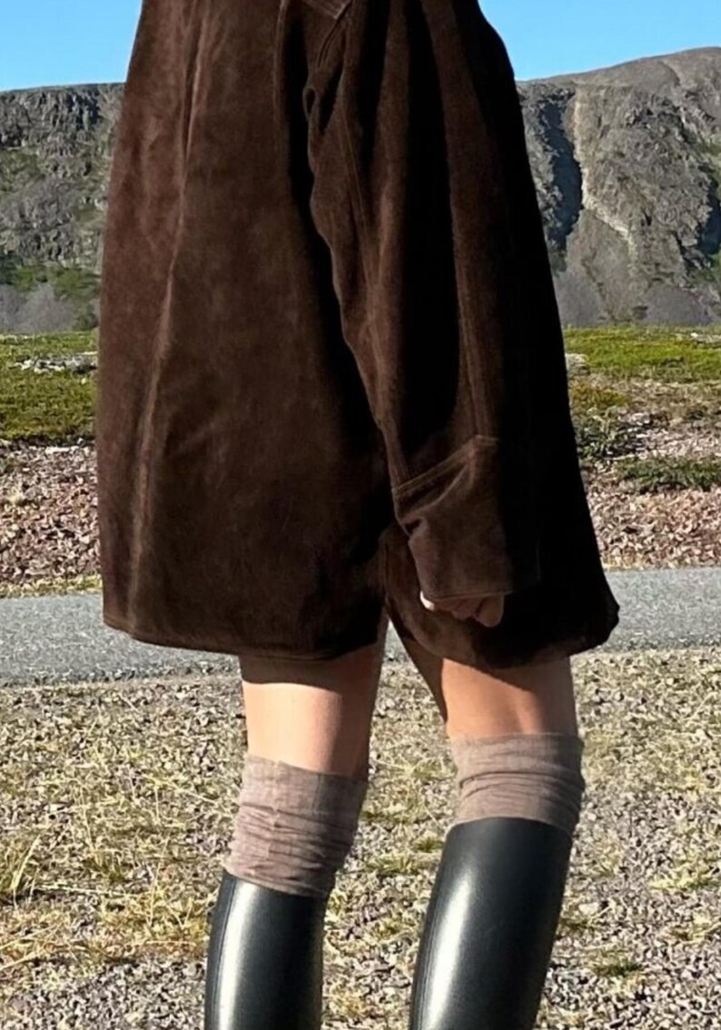 May include: A person wearing a dark brown suede jacket and matching shorts. The outfit is paired with gray socks and black rubber boots. The background features a mountain range and a paved path.