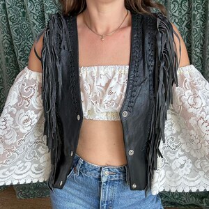 May include: A black leather vest with fringe detailing, braided edges, and snap closures. The vest is worn over a white lace crop top and paired with blue denim jeans. The sleeves are white lace.