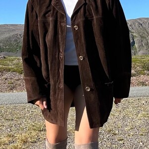 May include: A dark brown suede jacket with a collared neckline and button closures. The jacket has long sleeves and pockets. The person is wearing a white top, black shorts, gray socks, and black boots. The background features a mountain range and blue sky.