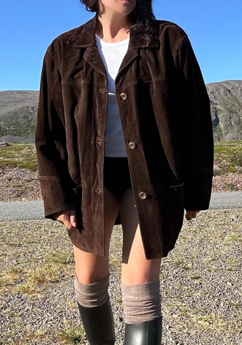 May include: A dark brown suede jacket with a collared neckline and button closures. The jacket has long sleeves and pockets. The person is wearing a white top, black shorts, gray socks, and black boots. The background features a mountain range and blue sky.