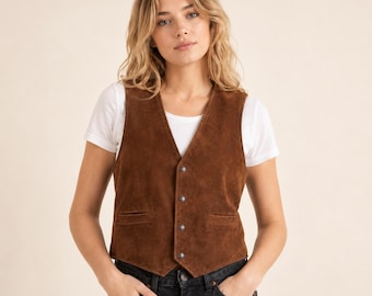 May include: A brown suede vest featuring a V-neckline, four silver-toned buttons down the front, and two horizontal welt pockets at the waist. The vest is worn over a plain white short-sleeved t-shirt and paired with dark denim jeans.