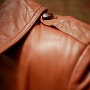 May include: Close-up of a brown leather jacket collar with a button closure. The leather has a smooth, slightly glossy finish and is stitched along the edges. The button is a dark, rounded shape.