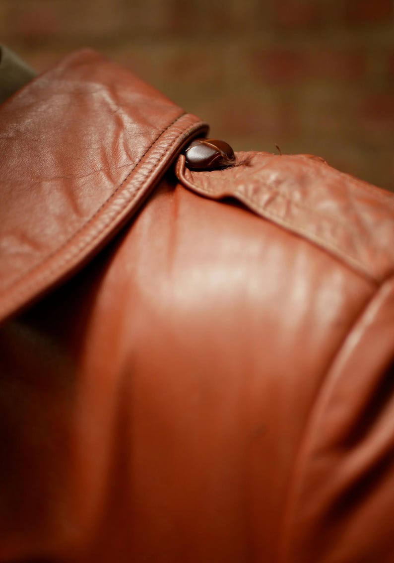 May include: Close-up of a brown leather jacket collar with a button closure. The leather has a smooth, slightly glossy finish and is stitched along the edges. The button is a dark, rounded shape.