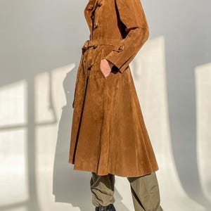 May include: A long, brown suede coat with a belted waist and button details. The coat has a flared skirt and a classic trench coat design. The model is wearing olive green pants and black high-heeled boots.