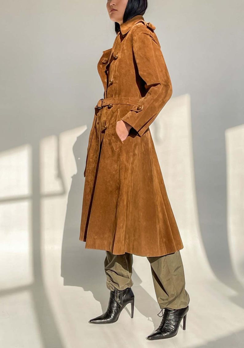 May include: A long, brown suede coat with a belted waist and button details. The coat has a flared skirt and a classic trench coat design. The model is wearing olive green pants and black high-heeled boots.