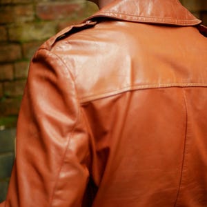 May include: A close-up of a brown leather jacket. The jacket has a collar, shoulder epaulets, and a back yoke. The leather appears to be smooth and has a slight sheen. The background is a blurred brick wall.