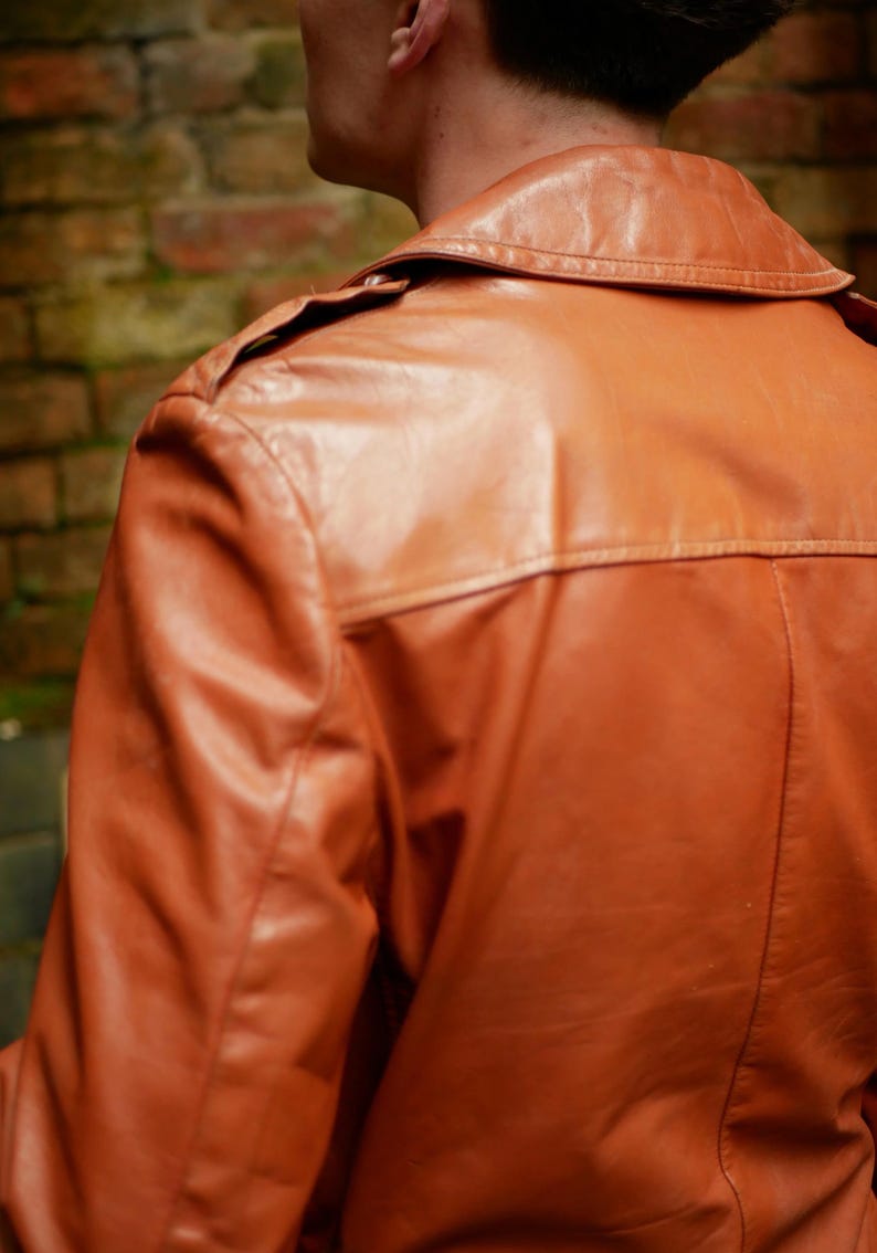 May include: A close-up of a brown leather jacket. The jacket has a collar, shoulder epaulets, and a back yoke. The leather appears to be smooth and has a slight sheen. The background is a blurred brick wall.