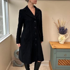 May include: A long, black coat with a classic design. The coat has a notched collar, button-down front, and long sleeves. The coat is knee-length and appears to be made of a wool or wool-blend fabric. The coat is worn with black boots.