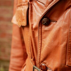 May include: Close-up of a vintage, orange-brown leather jacket. The jacket features a notched collar, two flap pockets, and a belt with a rectangular buckle. The buttons are a dark brown color. The leather has a worn, textured appearance.