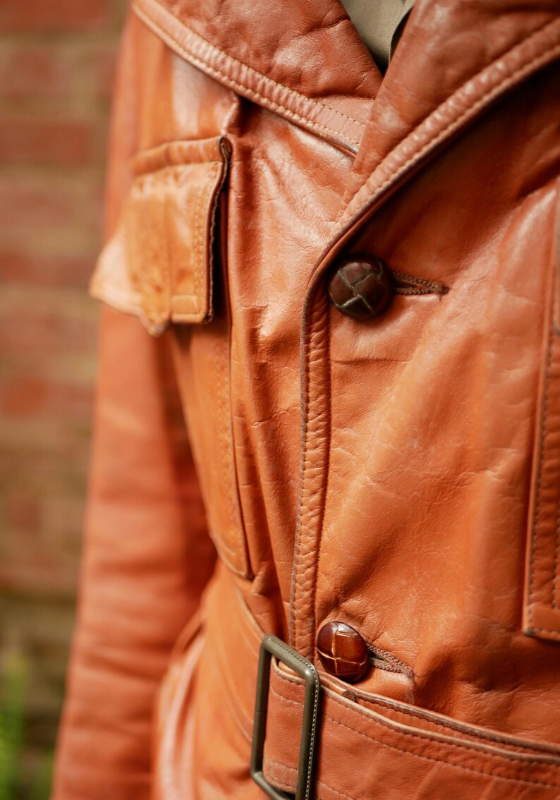 May include: Close-up of a vintage, orange-brown leather jacket. The jacket features a notched collar, two flap pockets, and a belt with a rectangular buckle. The buttons are a dark brown color. The leather has a worn, textured appearance.