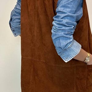 May include: A brown suede vest with a denim shirt underneath. The vest has a classic design with visible seams and a pocket. The denim shirt has long sleeves and buttoned cuffs. A gold watch is visible on the wrist.