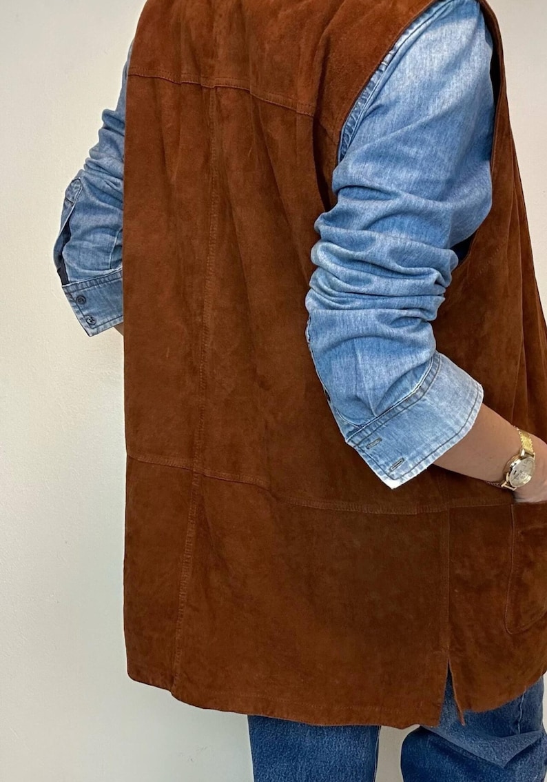 May include: A brown suede vest with a denim shirt underneath. The vest has a classic design with visible seams and a pocket. The denim shirt has long sleeves and buttoned cuffs. A gold watch is visible on the wrist.