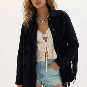May include: A black suede fringe jacket with long sleeves and a button-down front. The jacket is paired with a white top and denim shorts. The model is wearing layered necklaces.