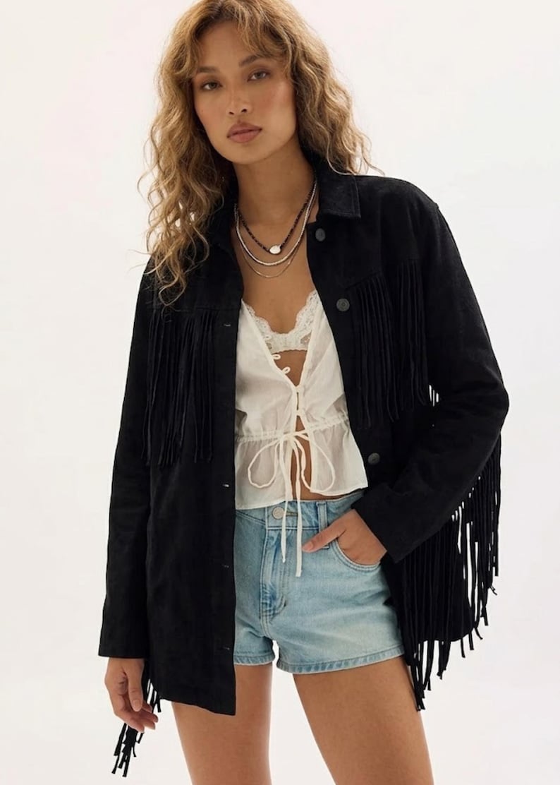May include: A black suede fringe jacket with long sleeves and a button-down front. The jacket is paired with a white top and denim shorts. The model is wearing layered necklaces.