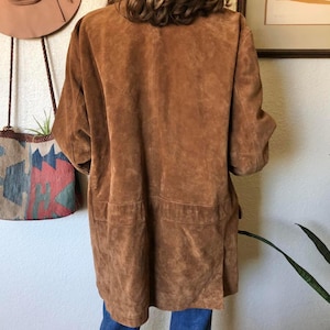 May include: A brown suede jacket with short sleeves and two front pockets. The jacket is knee-length and has a relaxed fit. The suede has a soft, textured appearance. The jacket is a warm, earthy tone.