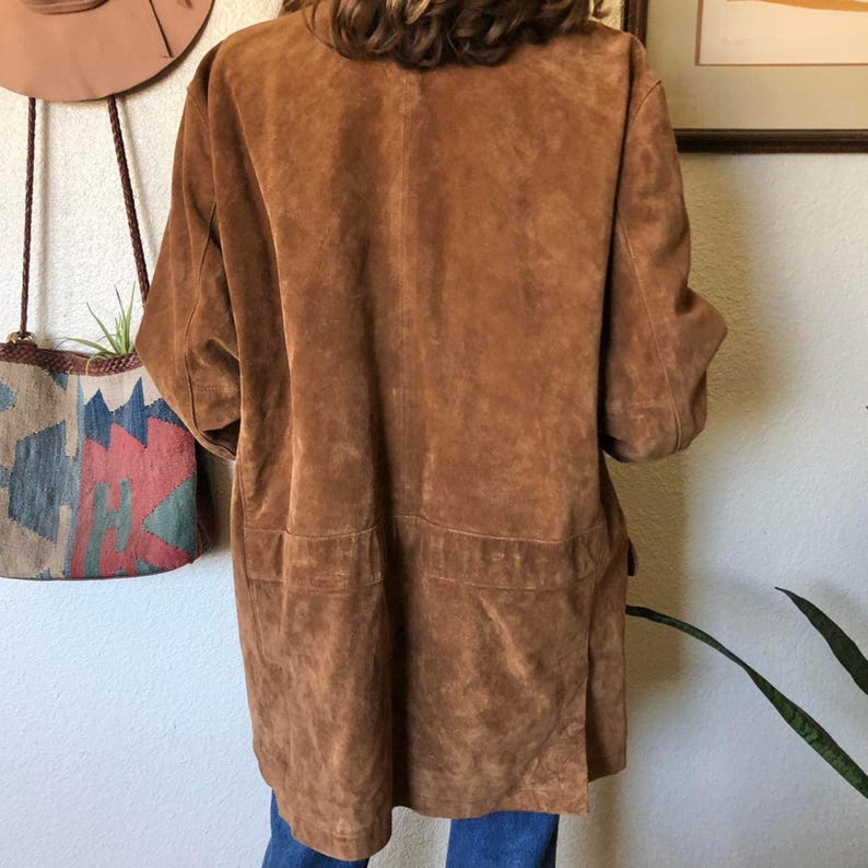 May include: A brown suede jacket with short sleeves and two front pockets. The jacket is knee-length and has a relaxed fit. The suede has a soft, textured appearance. The jacket is a warm, earthy tone.