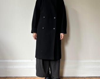 Puede incluir: Un abrigo largo negro de doble botonadura con cuello alto y seis botones oscuros. El abrigo llega a la rodilla y tiene mangas largas. La persona lleva pantalones grises oscuros y zapatos negros. El fondo es una pared lisa de color claro.