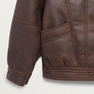 May include: A close-up of a brown leather jacket. The jacket features a classic design with a ribbed hem and cuffs, and a pocket. The leather has a distressed appearance, giving it a vintage look.