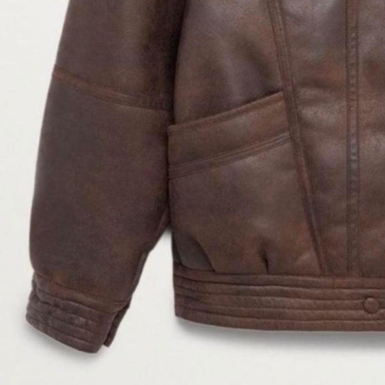 May include: A close-up of a brown leather jacket. The jacket features a classic design with a ribbed hem and cuffs, and a pocket. The leather has a distressed appearance, giving it a vintage look.