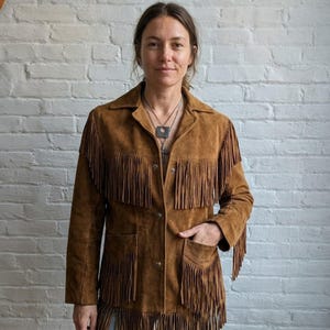 May include: A brown suede jacket with fringe detailing. The jacket features a collared neckline, button closures, and two front pockets. The fringe is along the sleeves, chest, and bottom hem. The jacket is worn by a person.