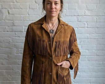 May include: A brown suede jacket with fringe detailing. The jacket has a collared neckline, button closures, and two front pockets. The fringe is along the sleeves, chest, and bottom hem. The jacket is worn by a person.