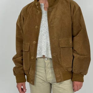 May include: A brown suede bomber jacket with a button-up front, two front pockets, and a ribbed collar and cuffs. The jacket is worn over a light-colored sweater and paired with khaki pants. The jacket has a classic, vintage style.