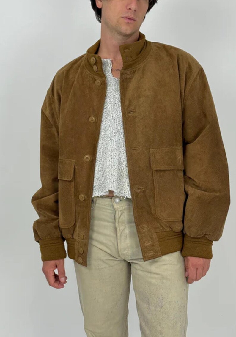 May include: A brown suede bomber jacket with a button-up front, two front pockets, and a ribbed collar and cuffs. The jacket is worn over a light-colored sweater and paired with khaki pants. The jacket has a classic, vintage style.