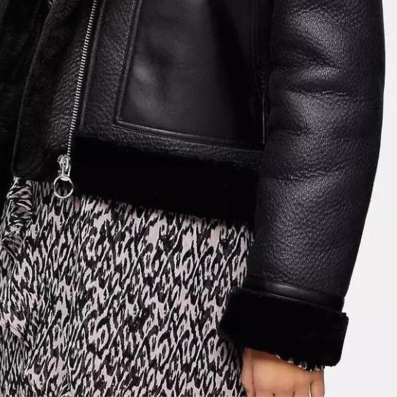 May include: Black leather jacket with a shearling lining and trim. The jacket features a zipper closure and a textured leather exterior. The sleeve cuff is trimmed with black shearling. The jacket is paired with a black and white patterned dress.
