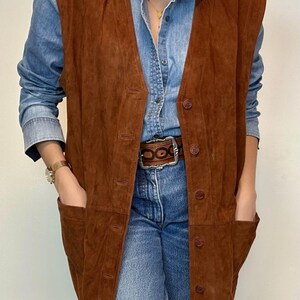 May include: A brown suede vest with a button-up front and two front pockets. The vest is worn over a denim shirt and blue jeans. The vest has a classic, vintage style.