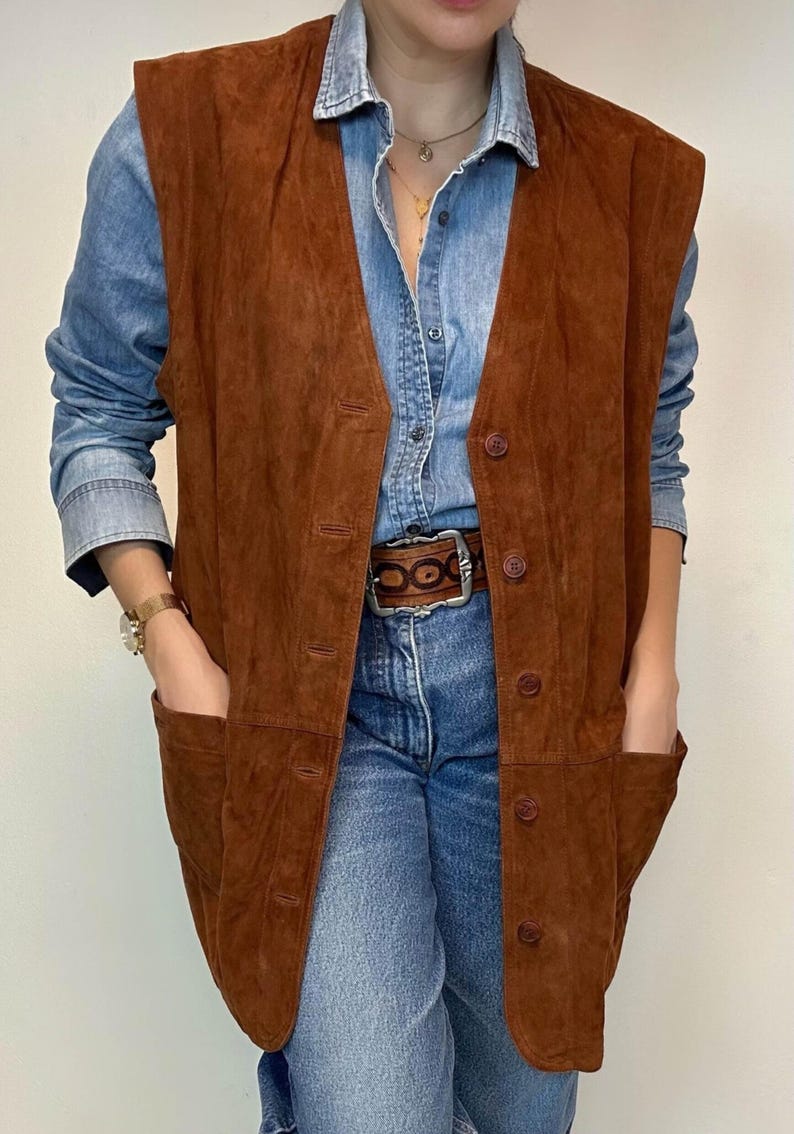 May include: A brown suede vest with a button-up front and two front pockets. The vest is worn over a denim shirt and blue jeans. The vest has a classic, vintage style.