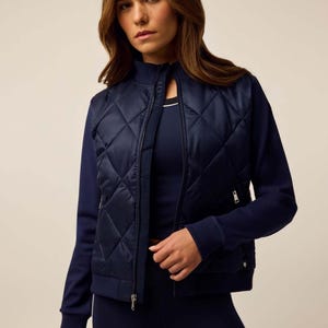 Puede incluir: Una chaqueta bomber acolchada de color azul marino con cuello alzado, mangas largas con puños acanalados y cremallera frontal completa. La chaqueta presenta un panel frontal acolchado con patrón de rombos y mangas lisas, sobre un top azul.
