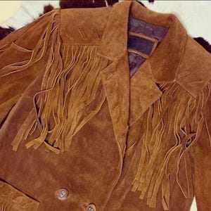 May include: A brown suede jacket with long fringe detailing on the chest and sleeves. The jacket has a classic collar and two button closure. The jacket is a warm brown color.