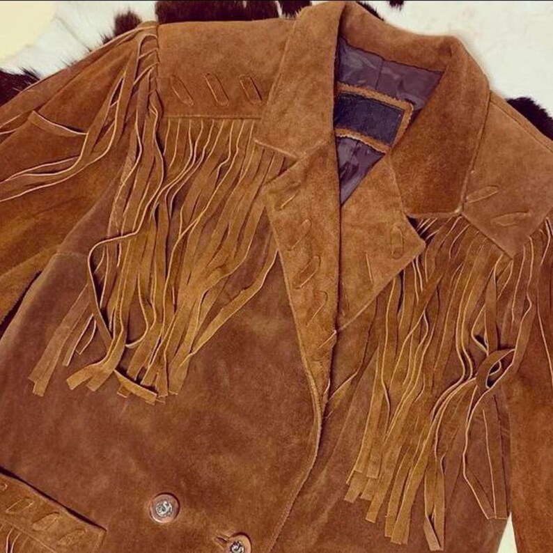 May include: A brown suede jacket with long fringe detailing on the chest and sleeves. The jacket has a classic collar and two button closure. The jacket is a warm brown color.