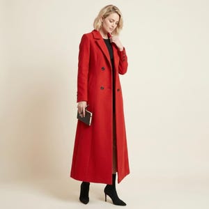 May include: A long, bright red double-breasted coat featuring a notched lapel, structured shoulders, and dark buttons down the front. The coat is worn over a black top and is paired with black ankle boots, while a small black clutch is held in hand.