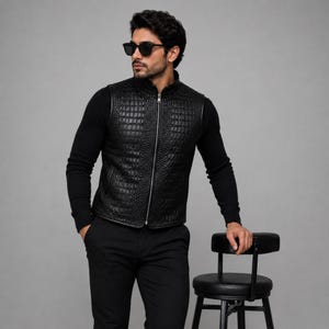 May include: A black sleeveless vest with a textured crocodile-skin pattern, featuring a full-length silver-tone front zipper, worn over a long-sleeved black shirt and black trousers, paired with dark-tinted sunglasses.