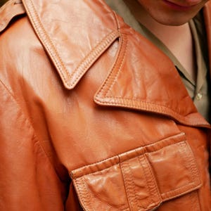 May include: Close-up of a vintage brown leather jacket. The jacket features a wide collar, two front pockets, and detailed stitching. The leather has a worn appearance, suggesting age and use. The jacket is part of a clothing collection.