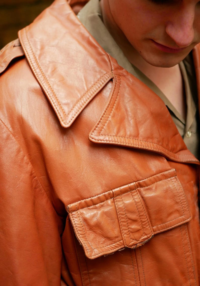 May include: Close-up of a vintage brown leather jacket. The jacket features a wide collar, two front pockets, and detailed stitching. The leather has a worn appearance, suggesting age and use. The jacket is part of a clothing collection.