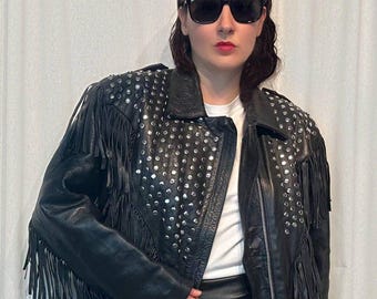 May include: Black leather jacket with silver studs and fringe detailing. The jacket has a classic biker style with a zipper closure and a notched collar. The jacket is worn with a white t-shirt and a black leather skirt. The person is wearing black sunglasses.