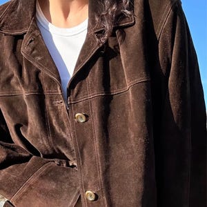May include: A brown suede jacket with a collared neckline and button closure. The jacket has a classic design with visible stitching details. The sleeves are long, and the jacket appears to be worn over a white t-shirt.
