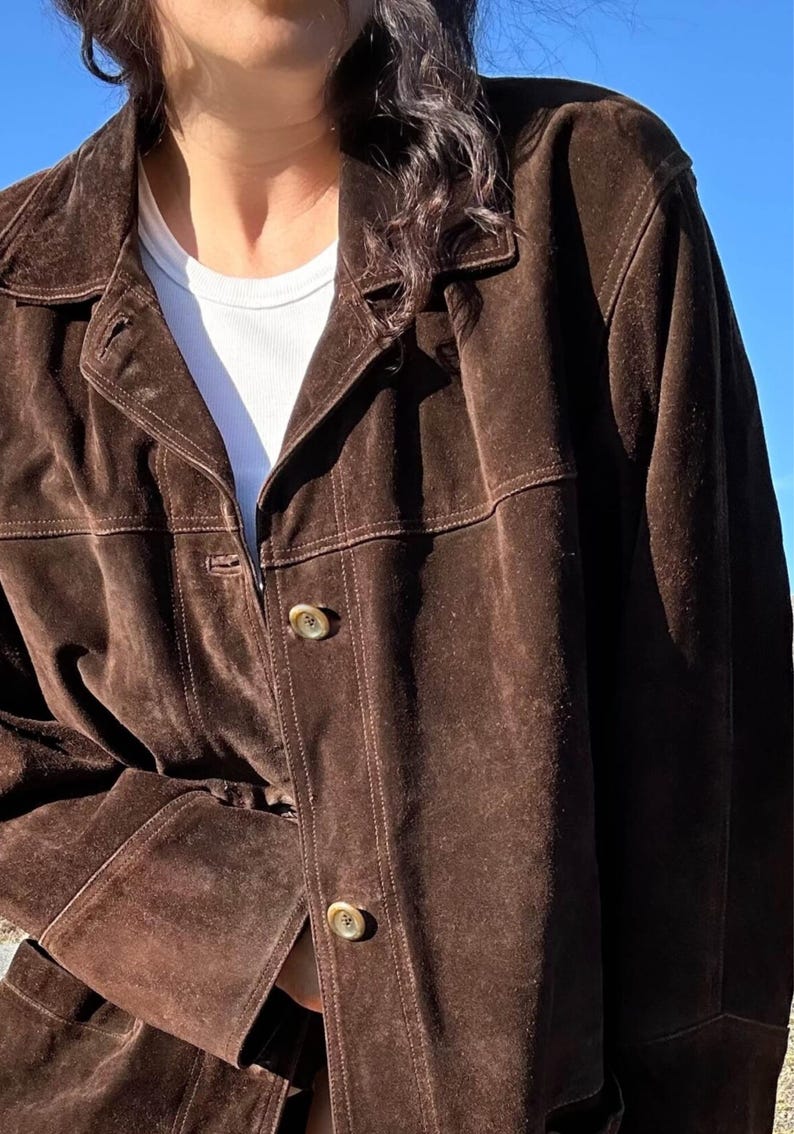 May include: A brown suede jacket with a collared neckline and button closure. The jacket has a classic design with visible stitching details. The sleeves are long, and the jacket appears to be worn over a white t-shirt.