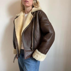 May include: A brown leather jacket with a cream-colored shearling lining and trim. The jacket features a classic asymmetrical zipper closure, a wide collar, and shearling cuffs. The jacket is a classic style, perfect for cold weather.